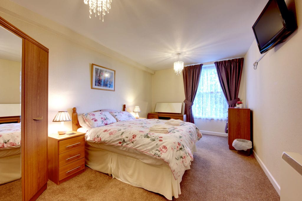 Lit bedroom containing a double bed with red flower covers plus wooden furniture and tv