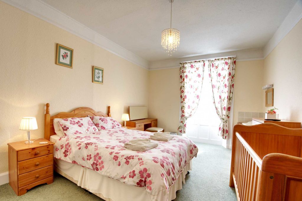 Light bedroom with double bed with red flower covers and wooden furniture including a cot