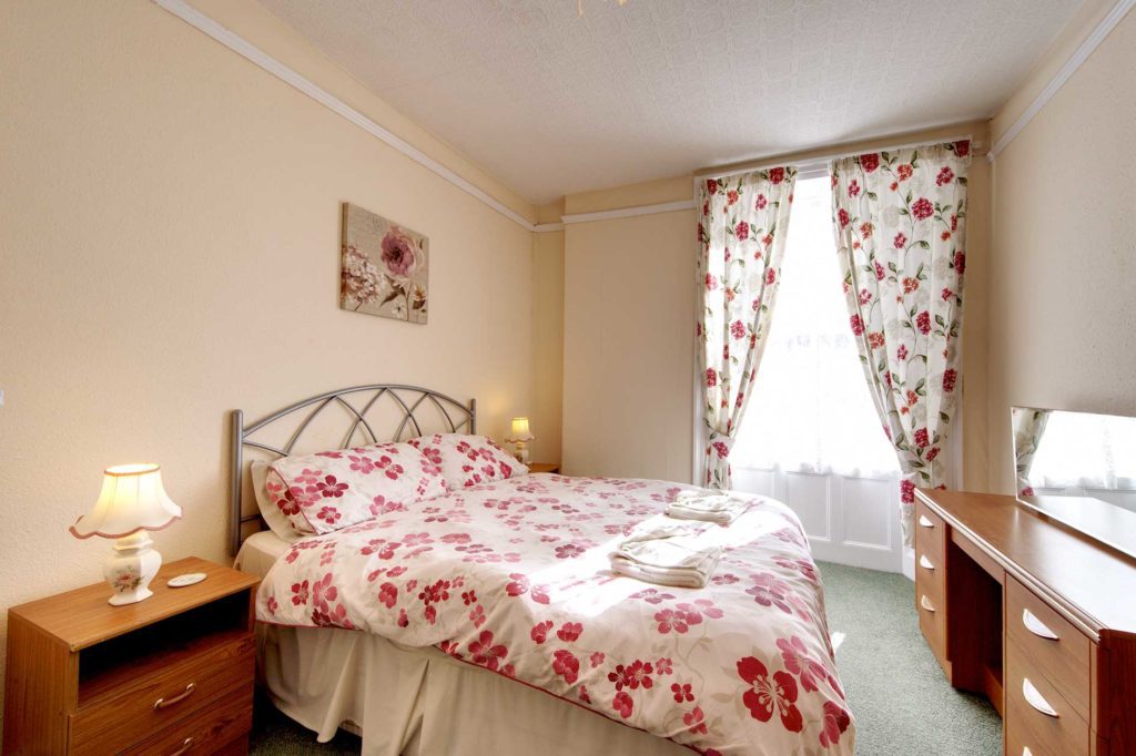 Light bedroom with double bed with red flower covers and wooden furniture