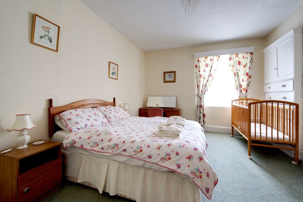 Light bedroom containing a double bed with red flower covers and curtains plus wooden furniture including a cot