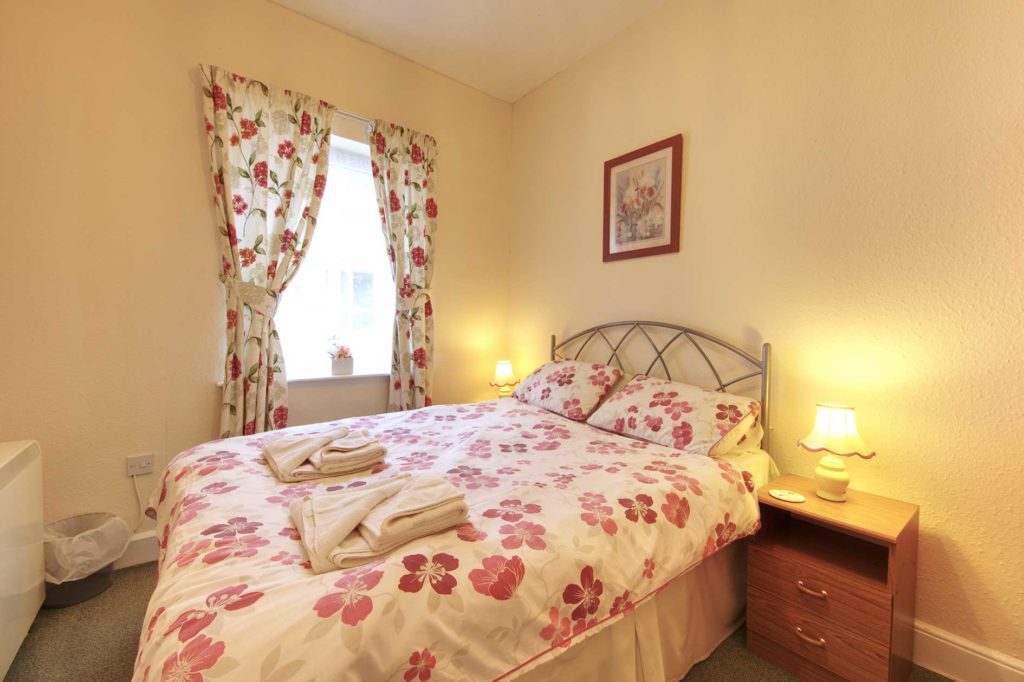 Lit bedroom containing a double bed with red flower covers and curtains plus wooden furniture